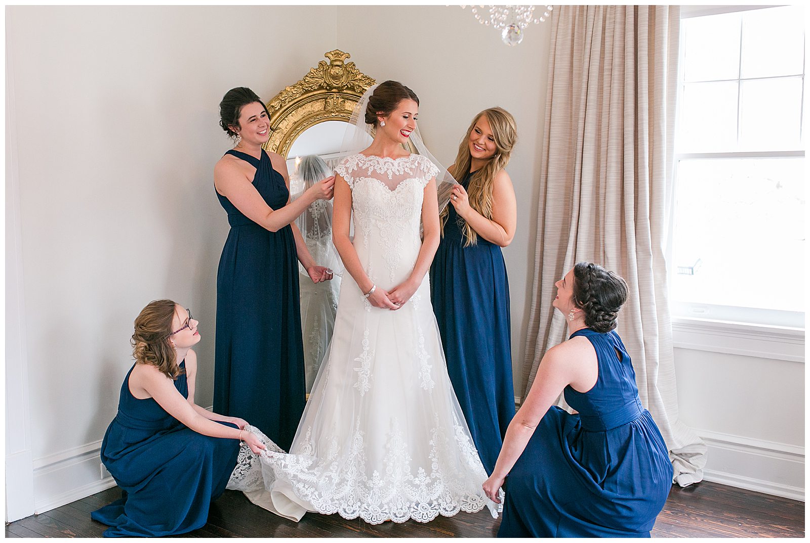bride has gown fixed by bridesmaids in front of full size gilded mirror