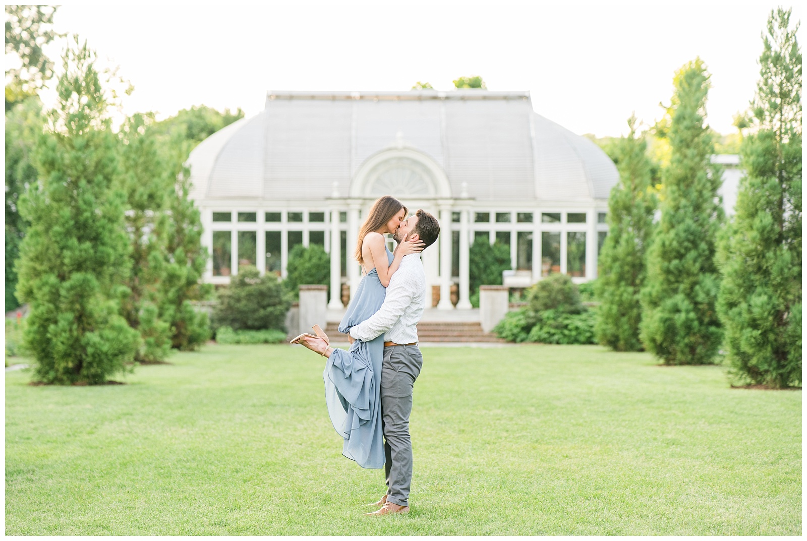 engaged couple dressed up in dusty blue flowing dress and button up shirt with grey dress pants poses romantically in front of a greenhouse in north carolina