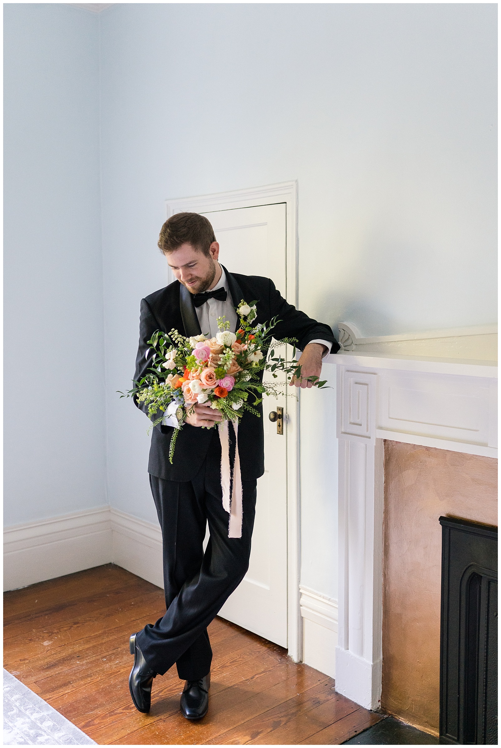 groom leans up against mantle and casually holds bridal bouquet with ribbon flowing down