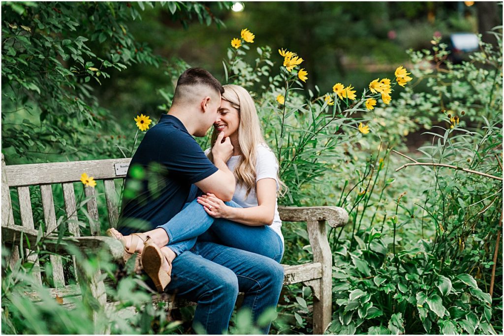 Couple sitting on a bench in a garden nuzzling, taken by an engagement photographer