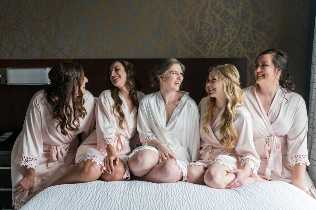 The bride and her bridesmaids sitting on a hotel bed laughing at each other during getting ready. during their Wedding in Raleigh NC