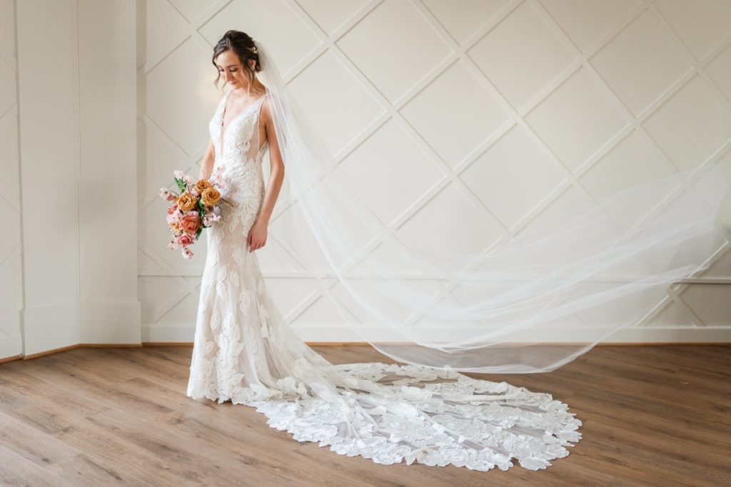 A woman standing in front of a white accent wall in a Magnolia Bride wedding gown with her veil flying.
