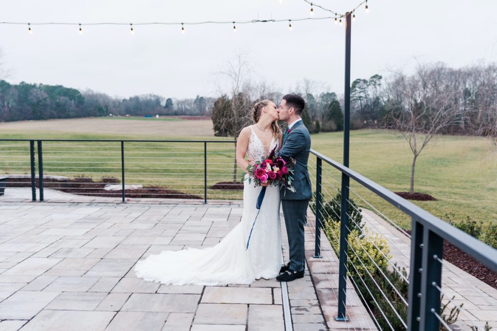 The bride and groom kissing on the back patio of Board and Batten over looking a green field.