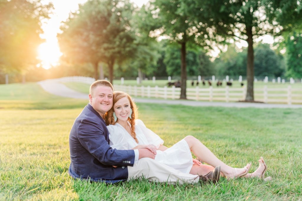A woman sitting in her fiance's lap as they sit in a field smiling during their engagement session.