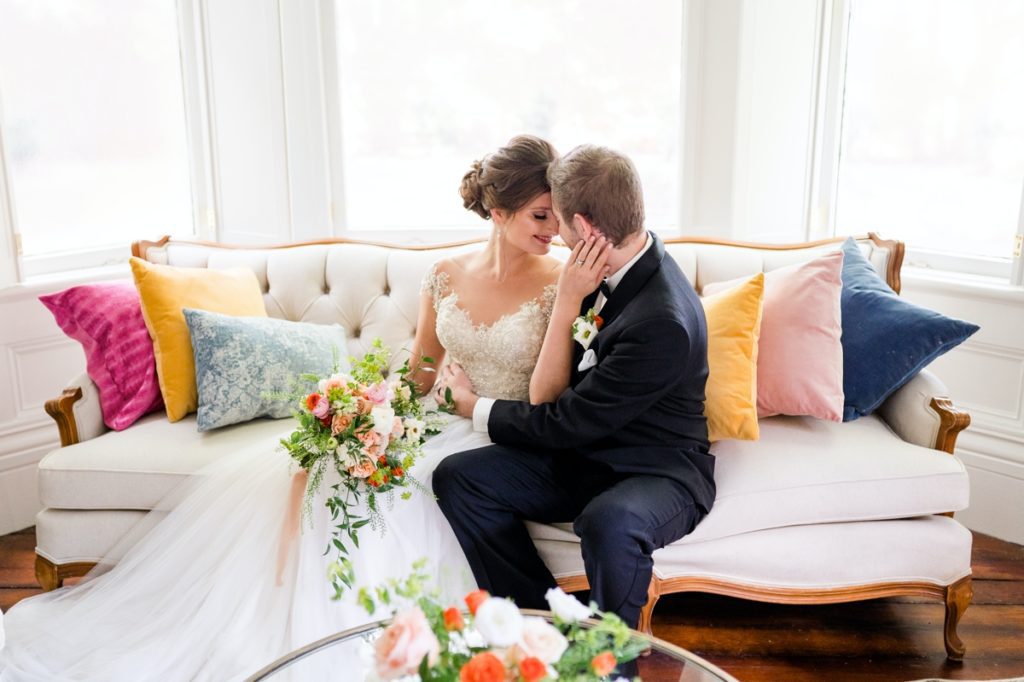 The bride and groom snuggling while she has one hand around the back of his head on a couch in a brightly lit getting ready room at Holt House.