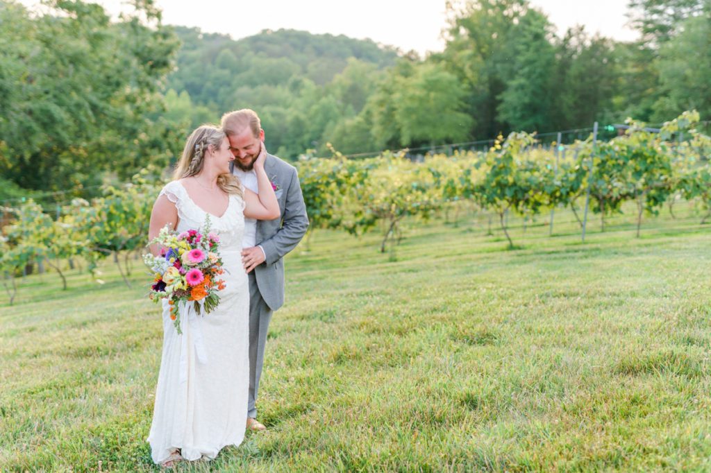 The groom stands behind the bride while she snuggles into his chest in a vineyard.