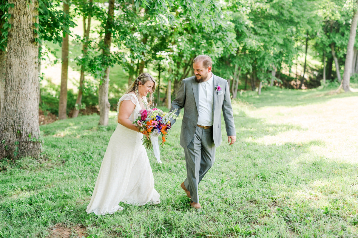groom leads his bride across a meadow in winston-salem, nc as she looks down at her brightly colored, boho bouquet