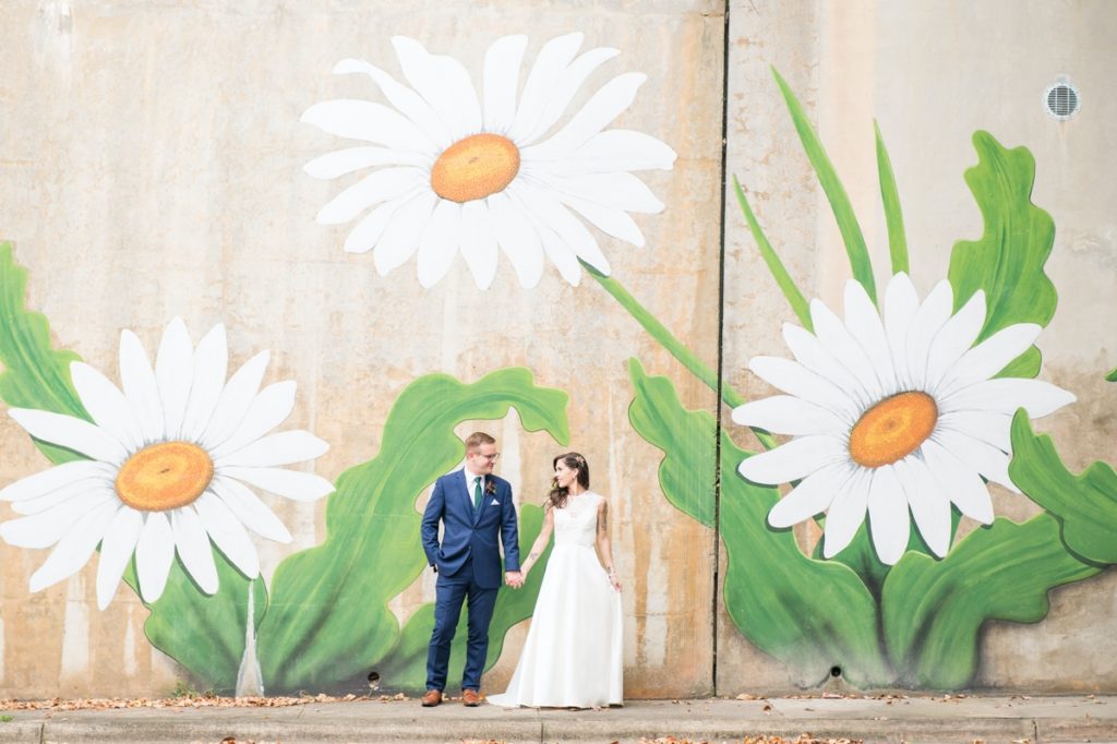 A man and woman holding hands in front of a daisy mural on their wedding day designed by Carly Marie Events