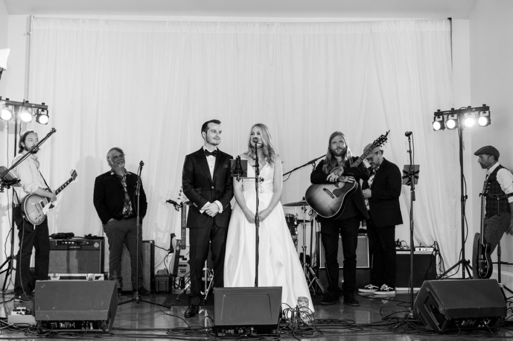The bride standing at a microphone saying a toast to her guests while the band waits behind. 