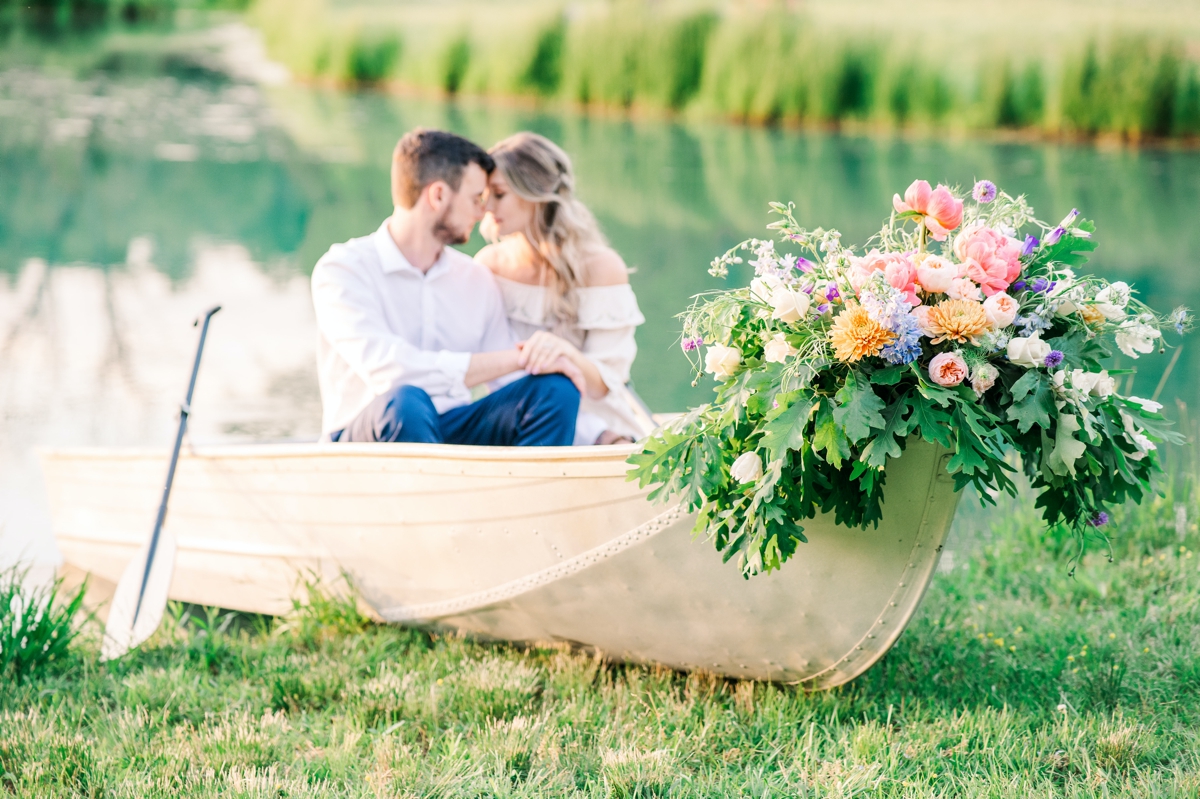 A man and woman sitting in a floral decorated canoe on the water's edge with their foreheads pressed together during their engagement session in the Triad area.