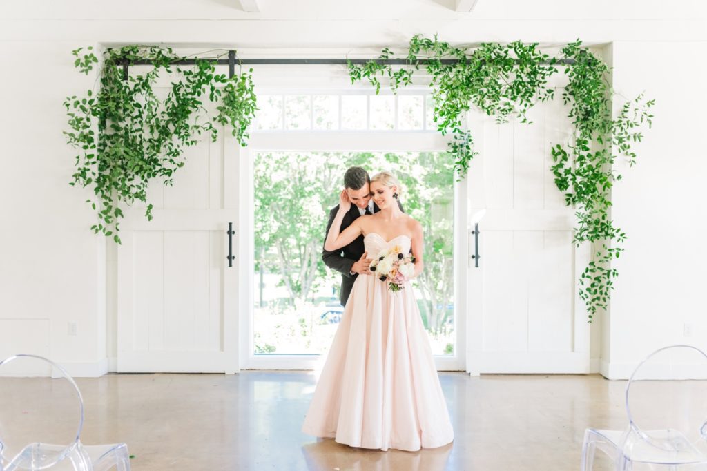 A groom standing behind his bride while he snuggles her in front of an ivy covered barn door during their wedding at The Barn at Reynolda Village.