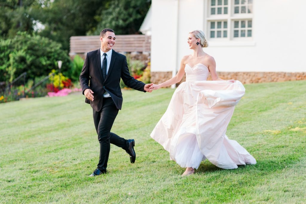 A bride and groom holding hands and smiling at each other while they run across the lawn of The Barn at Reynolda Village in Winston-Salem, NC.